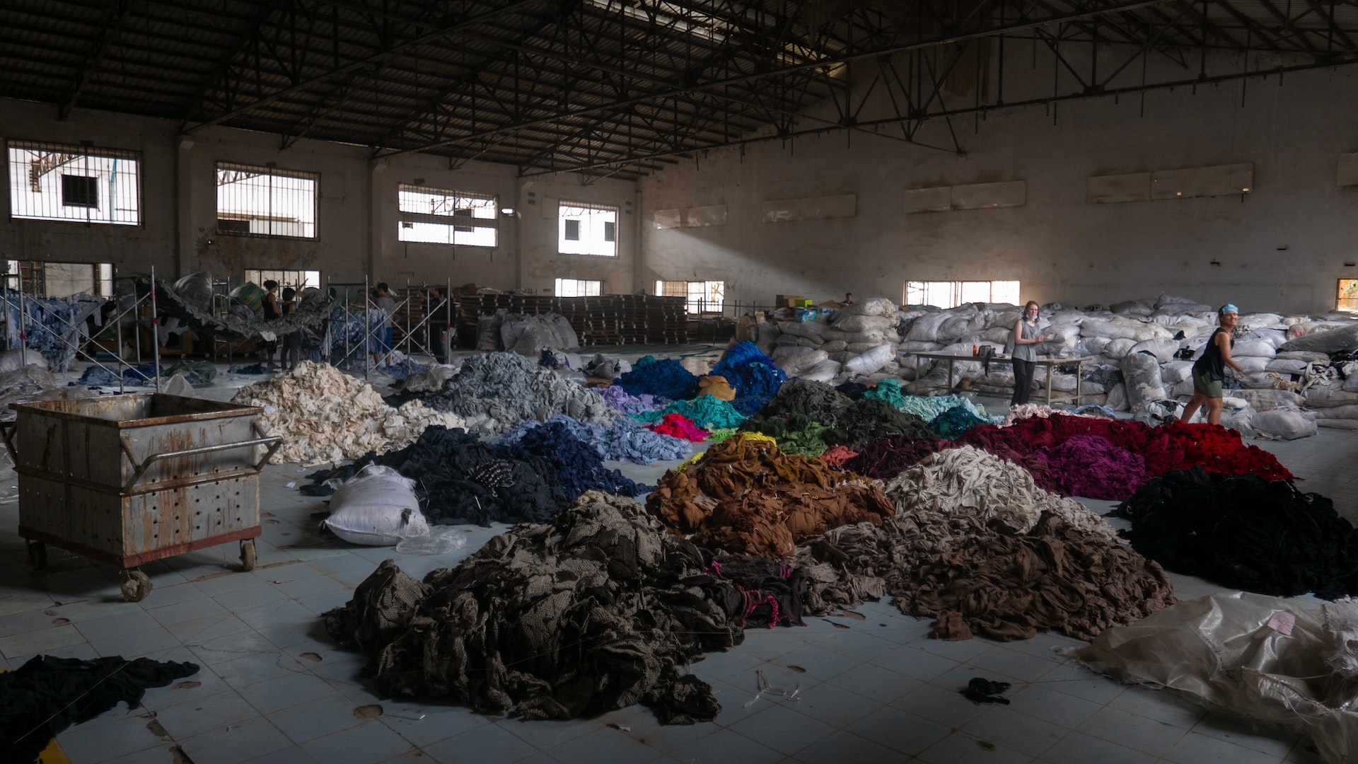 On the floor of a large and worn-out industrial hall, textile waste is arranged into piles based on their color.