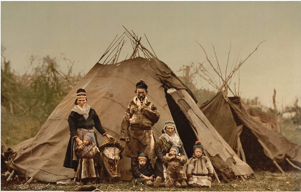 A Sámi family poses in front of two tents made of cloth and poles. Everyone has a skeptical expression.