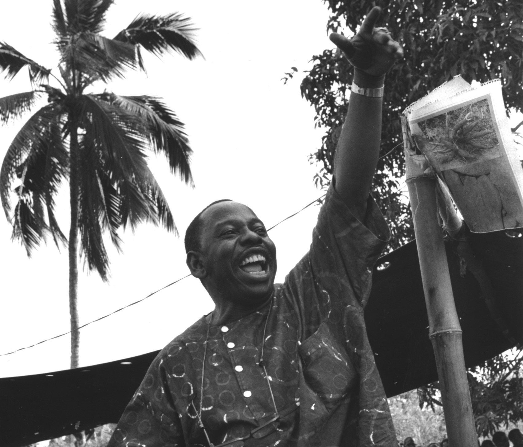 Joyful-looking Ken Saro-Wiwa is cheering. In the background, there is a canopy supported by bamboo sticks and a palm tree.