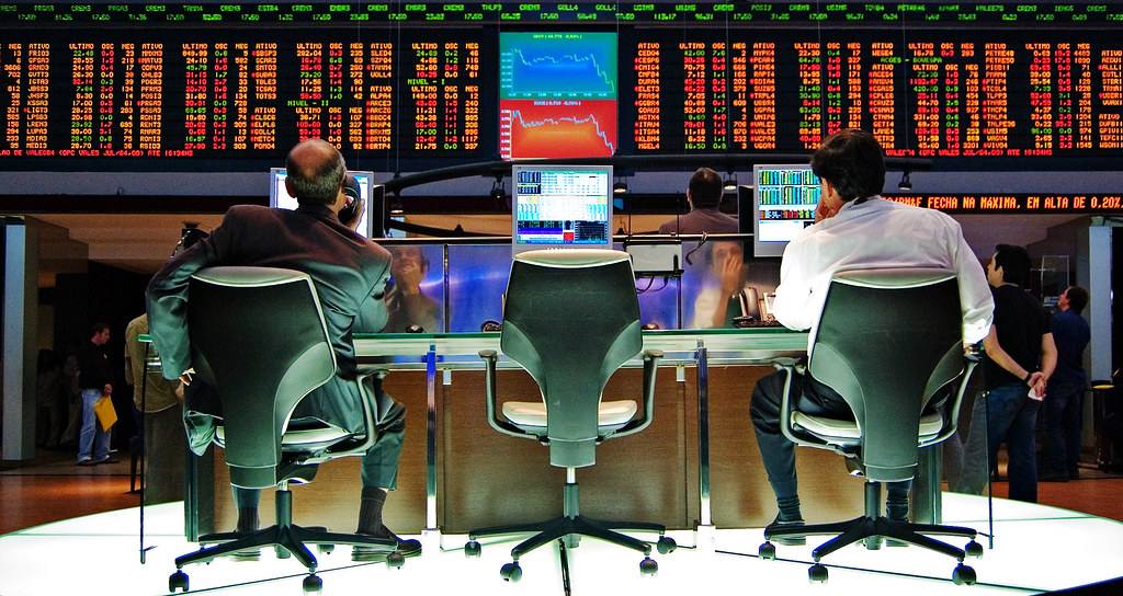 Individuals sitting in front of screens are watching plummeting stock market charts. Their expressions are concerned.